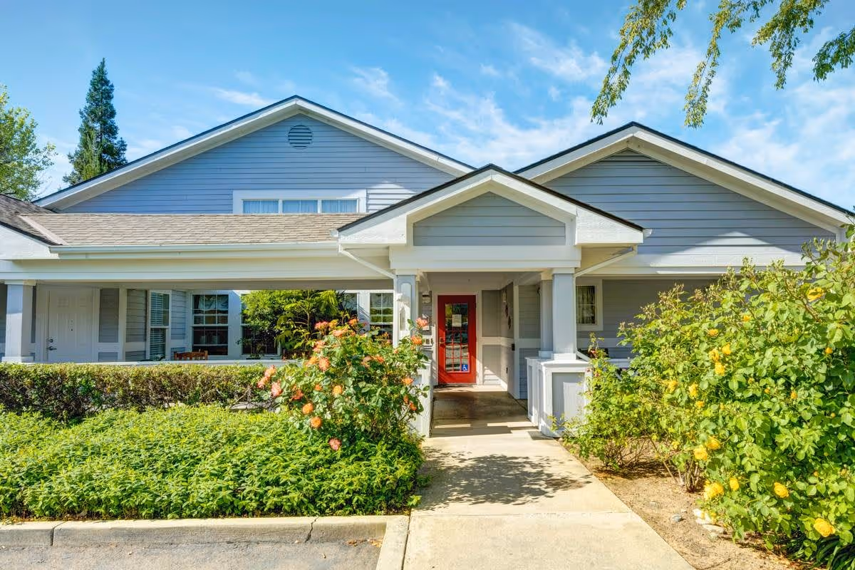 Exterior view of a single-story building with light blue siding and a red front door, surrounded by green bushes and flowering plants under a blue sky.