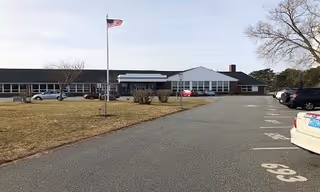 Single-story senior care facility with a parking lot, flagpole, and several parked cars.