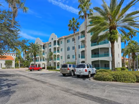 Mediterranean-style multi-story residential building with balconies, palm trees, and parked cars in the foreground.
