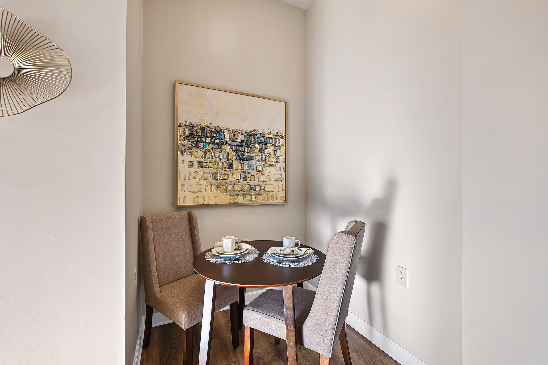 Small dining nook with round table, two upholstered chairs, cups, and wall art.