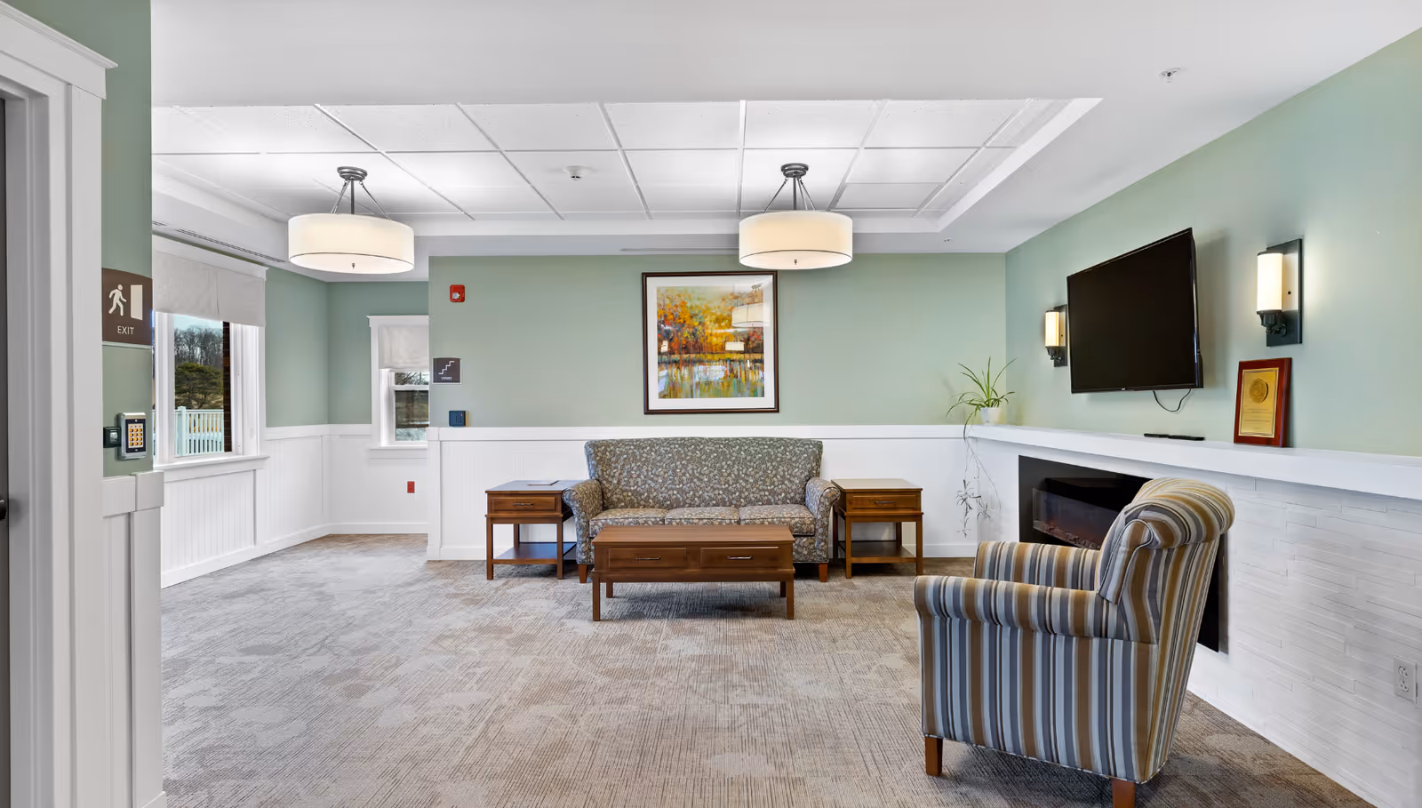 Bright senior living lounge with sofa, striped chair, coffee tables, TV, and fireplace.