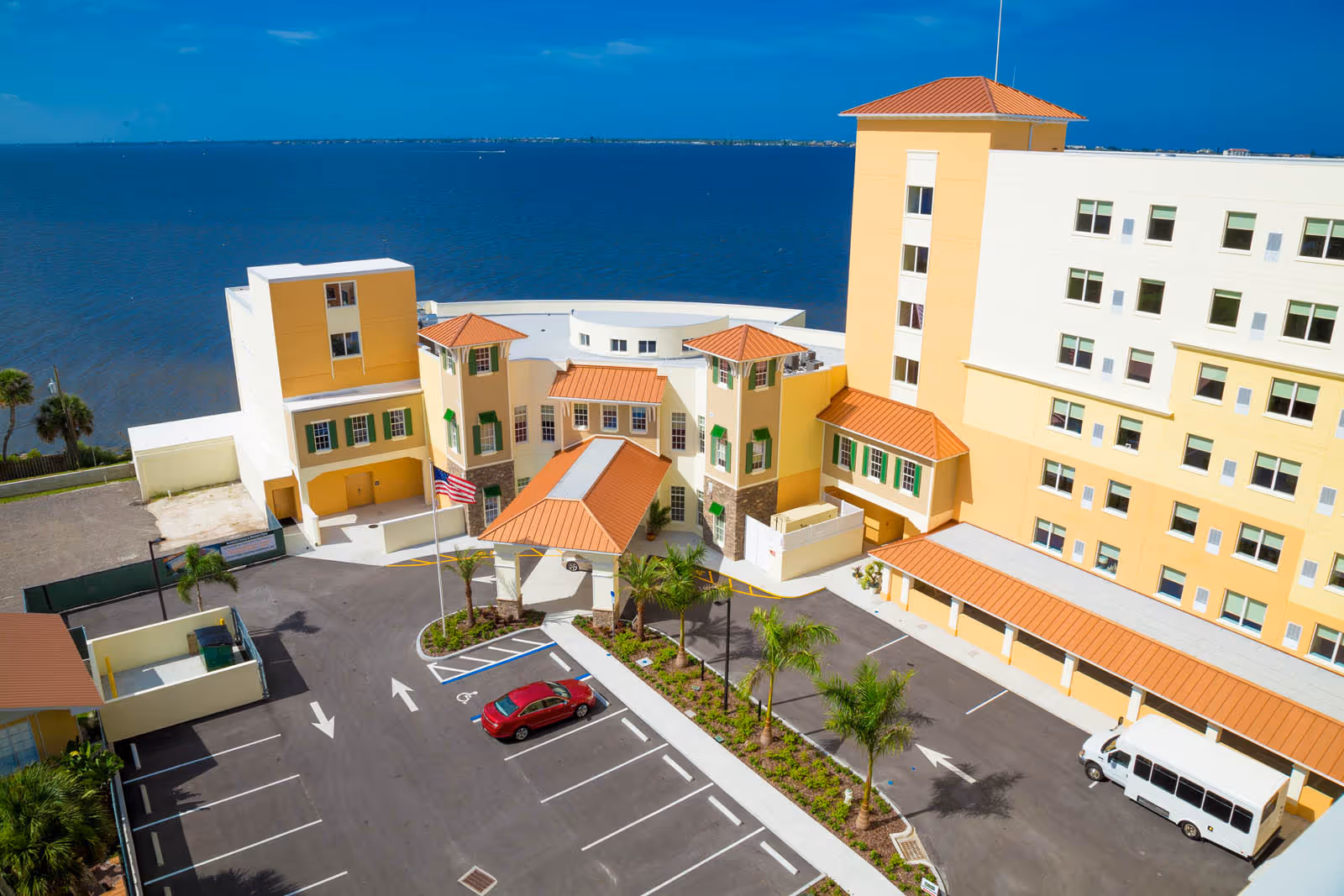 Aerial view of the Grand Villa of Palm Bay senior living facility showing a multi-story building with yellow and white exterior walls and orange tiled roofs. The facility is located next to a large body of water. There is a parking lot with a few vehicles, palm trees, and an American flag near the entrance.