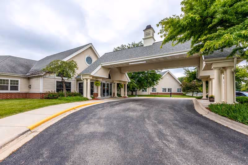 Covered porte-cochère and front entrance of a single-story care facility with columns, driveway, and landscaped grounds.