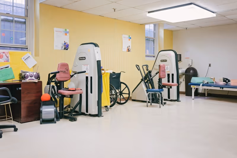 A rehab therapy room with exercise machines, a wheelchair, a desk, and a treatment table.