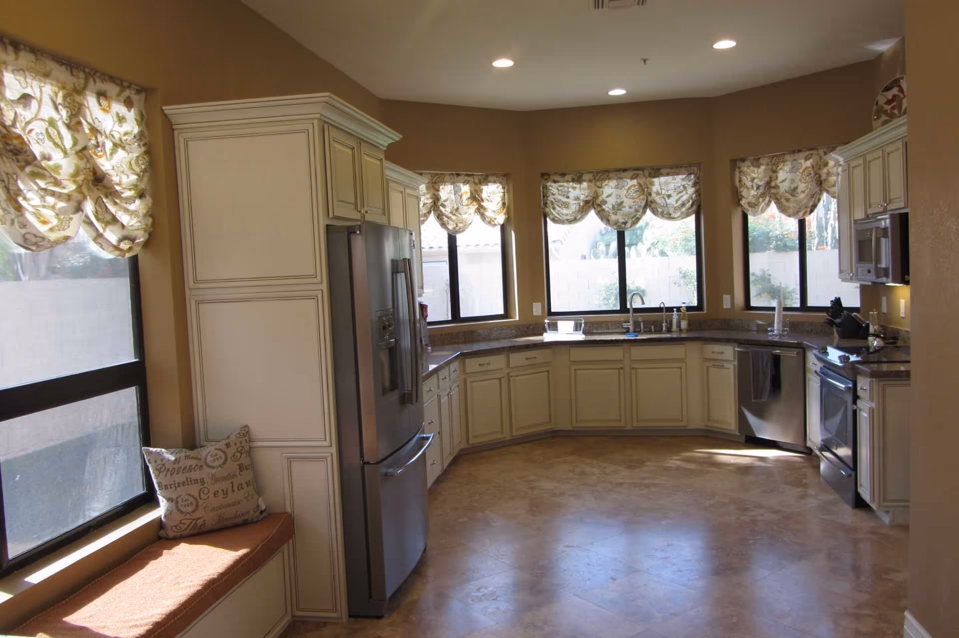 A spacious kitchen with cream-colored cabinets and granite countertops arranged in a U-shape. The kitchen features stainless steel appliances including a refrigerator, dishwasher, and oven. There are four windows with floral-patterned valances letting in natural light. A cushioned bench with decorative pillows is positioned under one window.