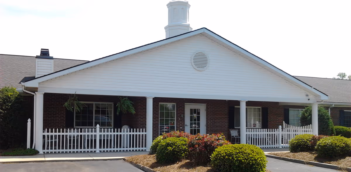 Front exterior view of a single-story brick building with white trim and a covered porch supported by white columns. The porch has white picket fencing and hanging plants. There are bushes and landscaping in front of the building.