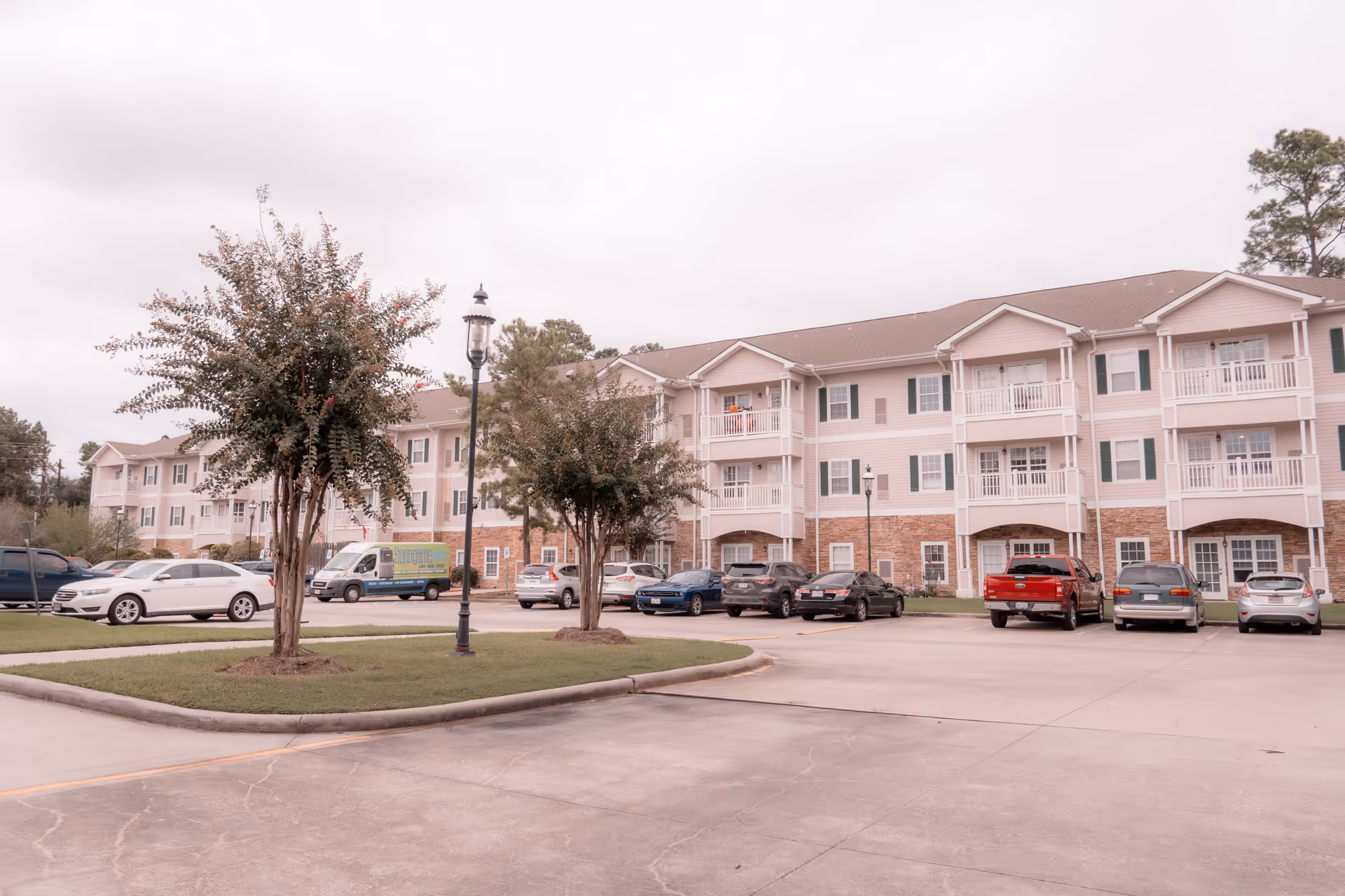 Exterior view of a three-story residential building with balconies, surrounded by a parking lot with several parked cars and small landscaped areas with trees and grass under a cloudy sky.