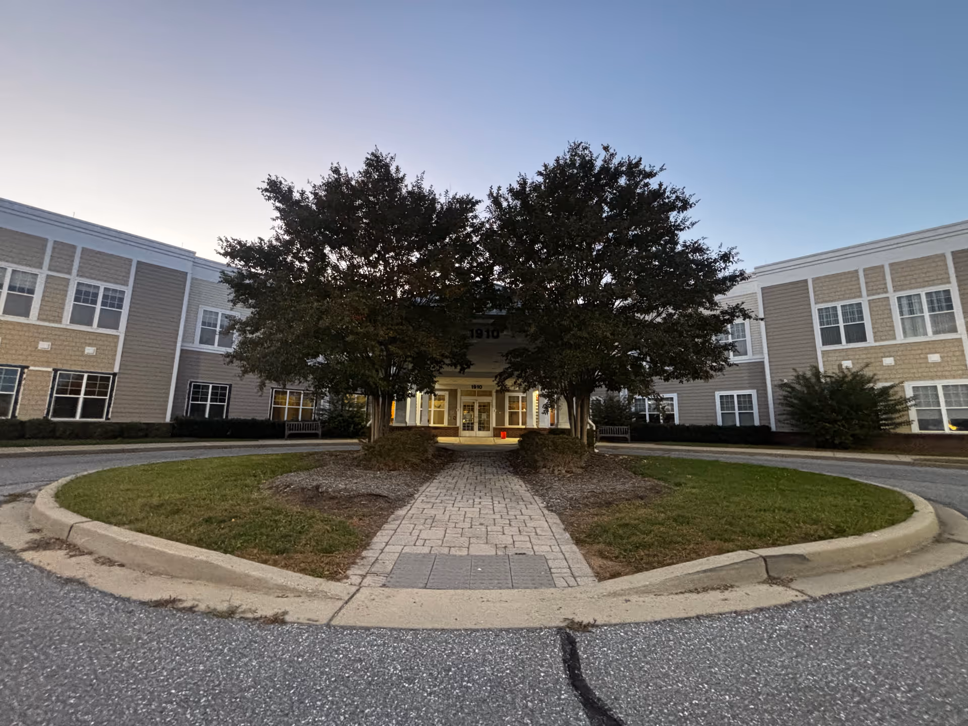 Front exterior view of Odyssey Assisted Living at Montevue building with a circular driveway, two large trees flanking the entrance, and a pathway leading to the main doors under a covered entryway.