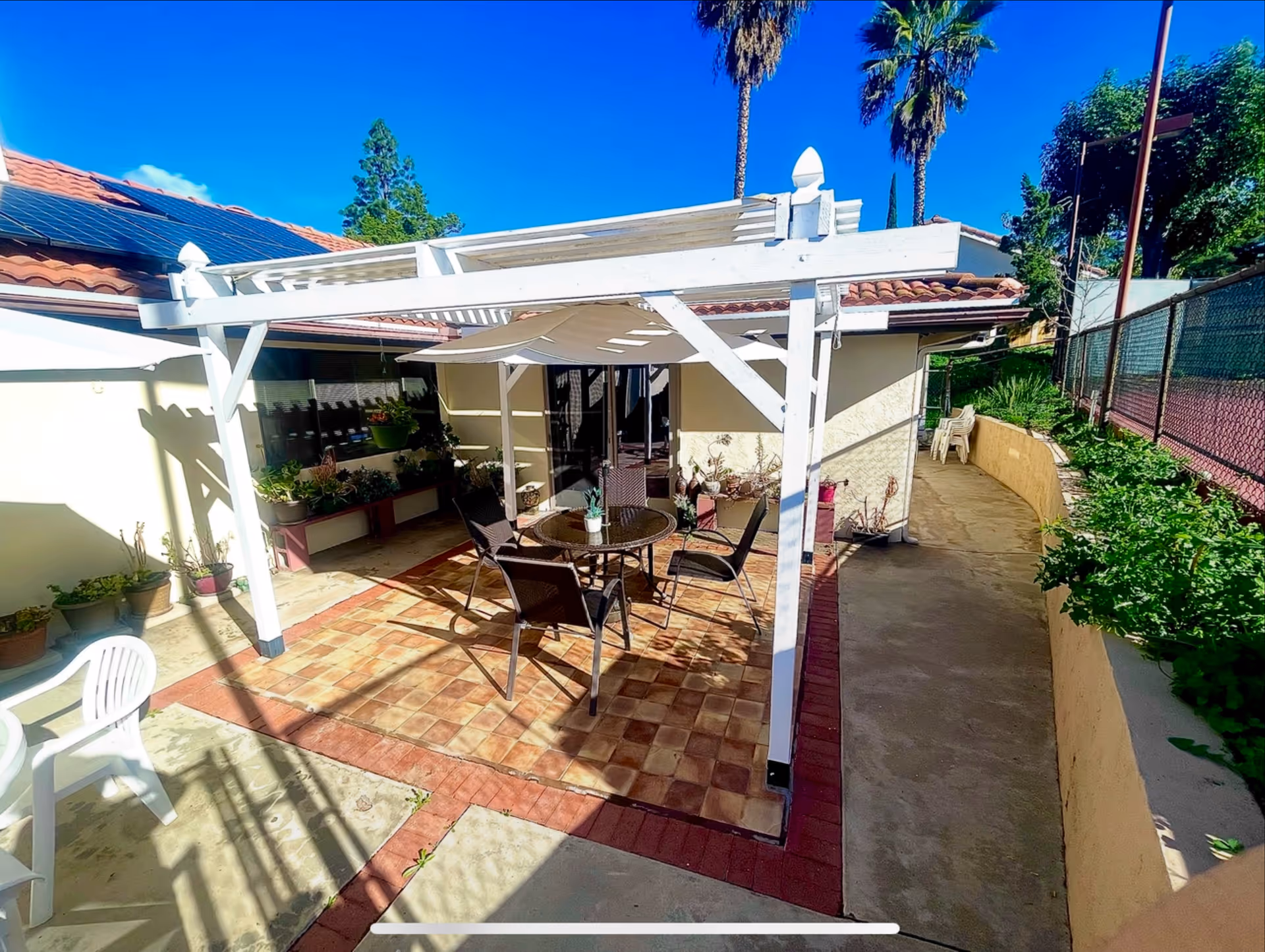 Outdoor patio area with a white pergola covering a round glass table and four chairs. There are potted plants along the walls and a concrete pathway beside the patio. Palm trees and a clear blue sky are visible in the background.