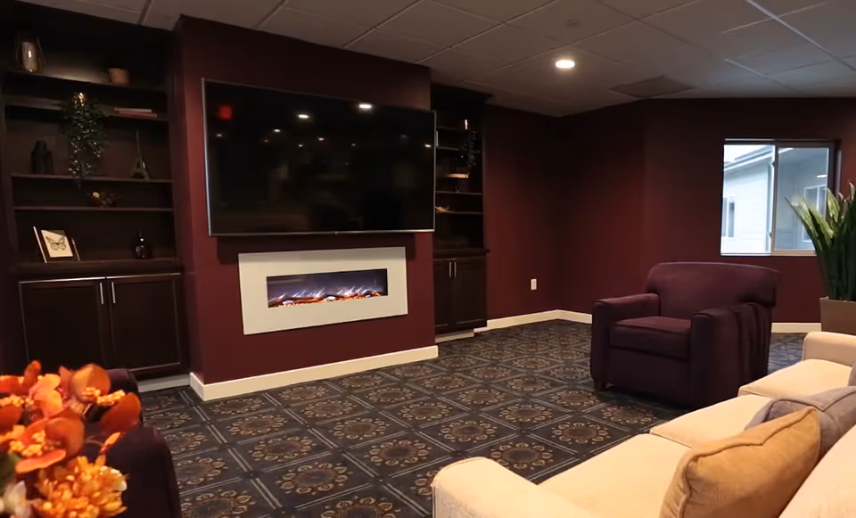 Cozy lounge with a wall-mounted TV above an electric fireplace, upholstered chairs and a sofa, built-in shelving, and patterned carpet.