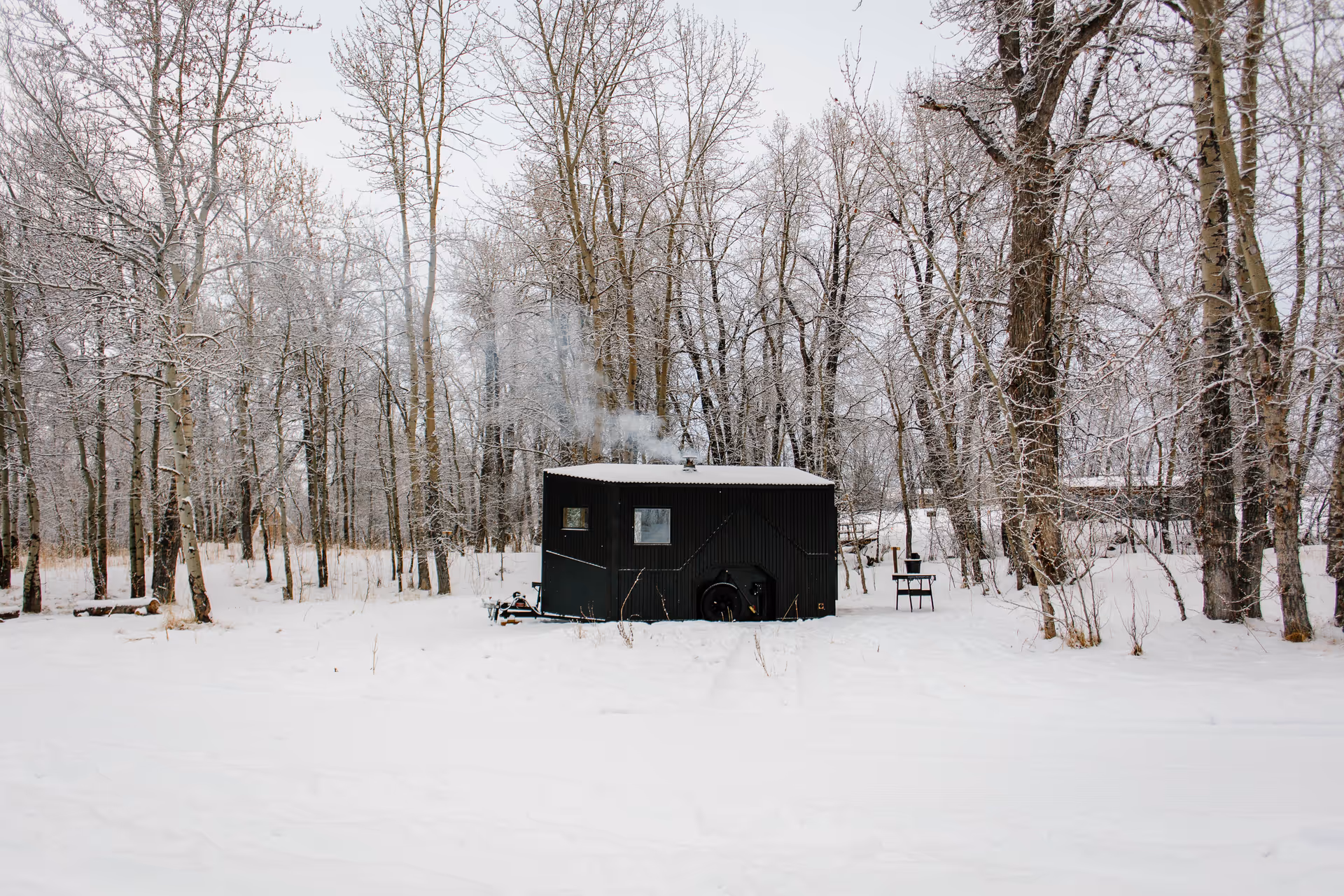 Black sauna cabin sits in a snow-covered forest, with smoke rising from its chimney.