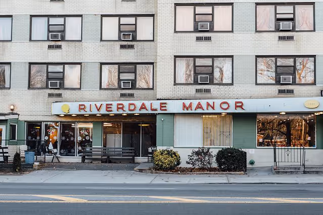 Front exterior of the Riverdale Manor building showing its street-level entrance, large sign, windows with air conditioners, benches, and small shrubs.