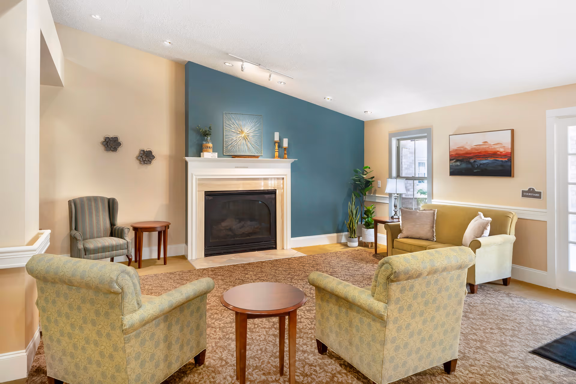 A cozy living room area with a fireplace centered on a blue accent wall. The room features two patterned armchairs facing a small round wooden table, a striped armchair next to a small side table, and a light green sofa with pillows near a window and a side table with a lamp. The walls are painted beige, and there is a carpet covering the floor. Decorative items include wall art above the fireplace and a painting on the wall near the sofa, along with some potted plants.