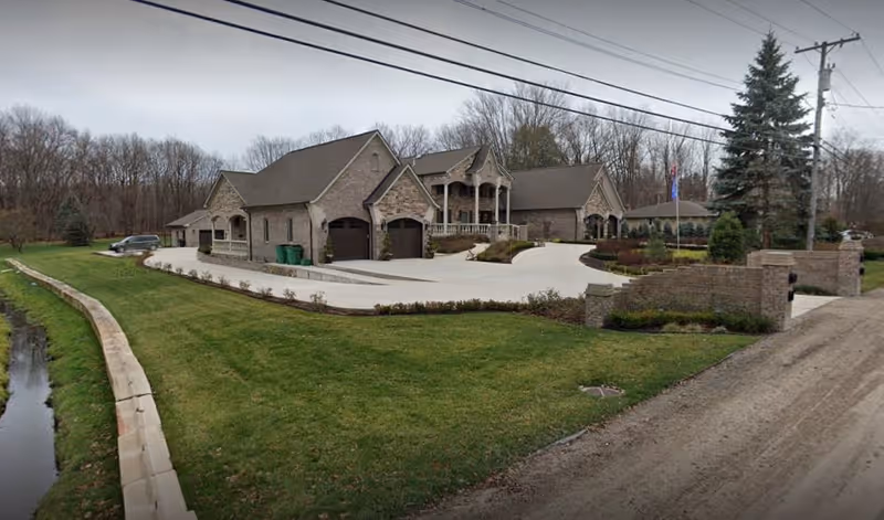 A large brick residential-style building with multiple gables and a covered porch, surrounded by a well-maintained lawn and landscaping. There is a paved driveway leading to two garage doors, and a dirt road runs alongside the property. Trees without leaves are visible in the background under a cloudy sky.