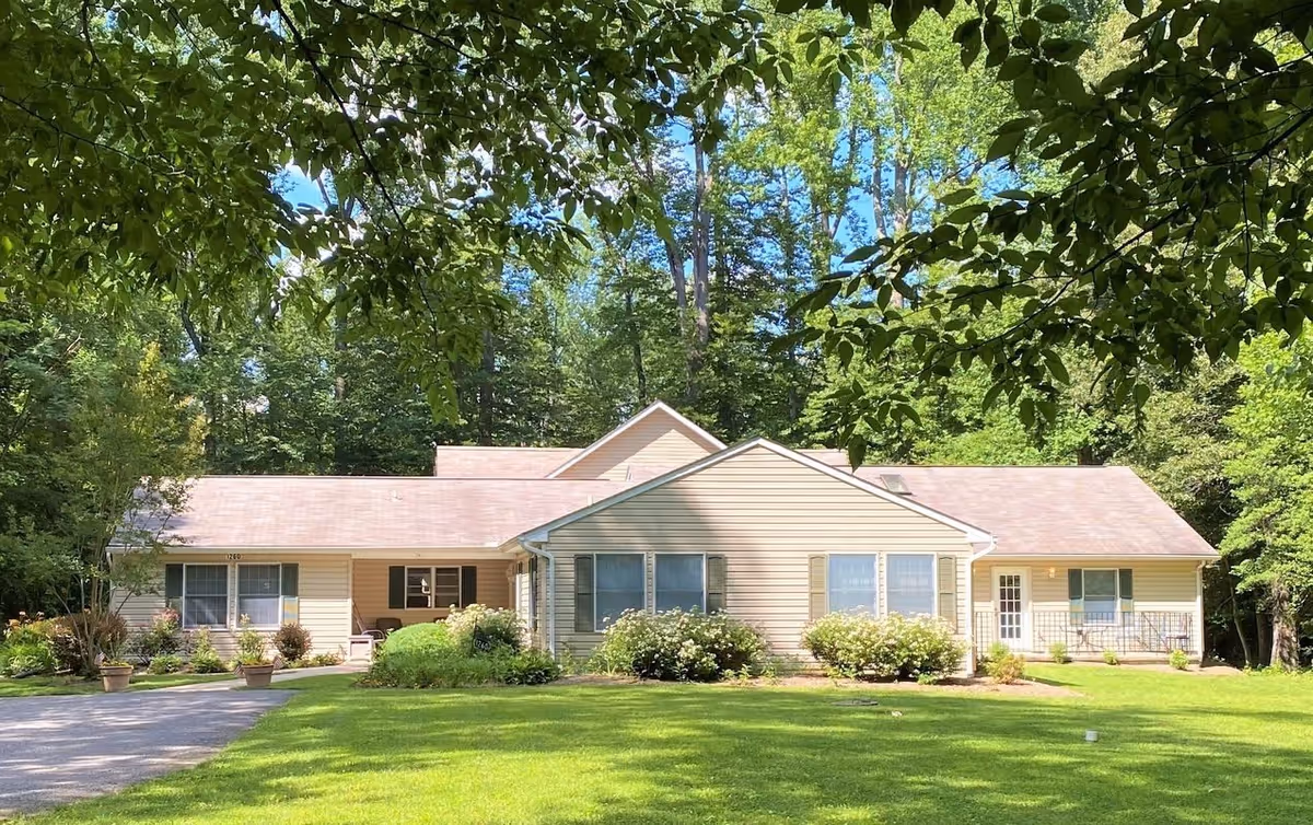 Front exterior of a single-story beige assisted living building with gabled roof, surrounded by lawn and trees.