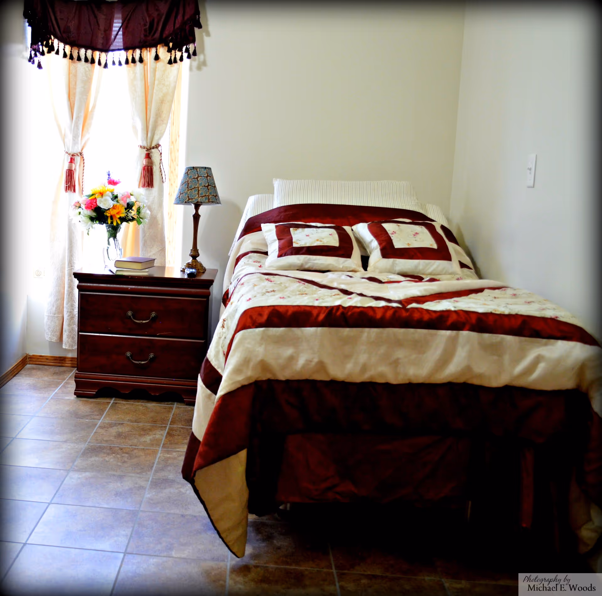 A neatly made bed with burgundy-cream quilt beside a wooden nightstand, lamp, and flowers.