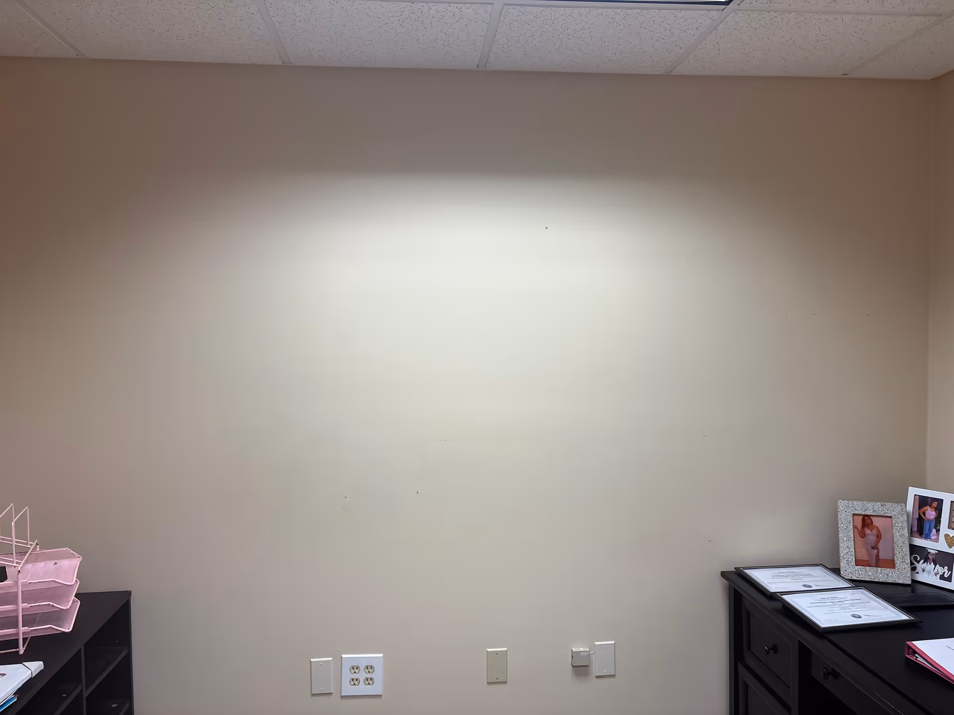 Blank beige wall in a small office corner with desk, framed photos, and storage.