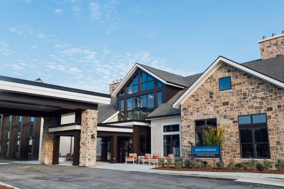 Exterior view of The Spires at Berry College showing the main entrance with stone and wood facade, large windows, a covered driveway, and outdoor seating under a clear blue sky.
