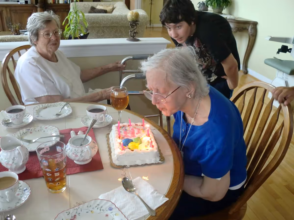 An elderly woman in a blue shirt is blowing out candles on a birthday cake at a dining table. Another elderly woman in a white shirt is sitting nearby, and a young person is leaning over to watch. The table is set with teacups, plates, and glasses, and the setting appears to be a cozy indoor living space.