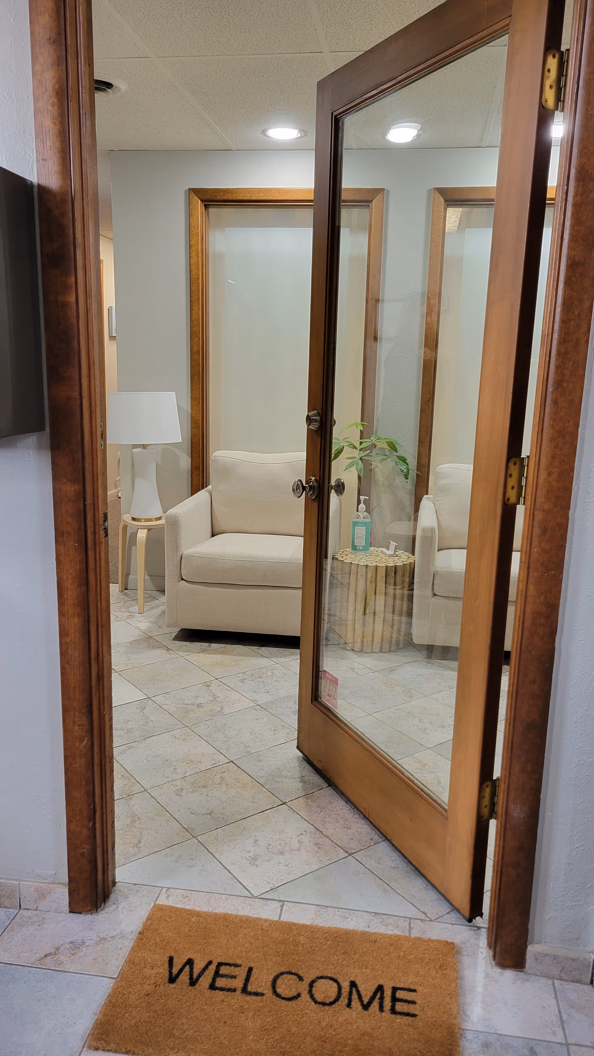 Glass door opens to a beige lounge area with plant, lamp, and welcome mat.