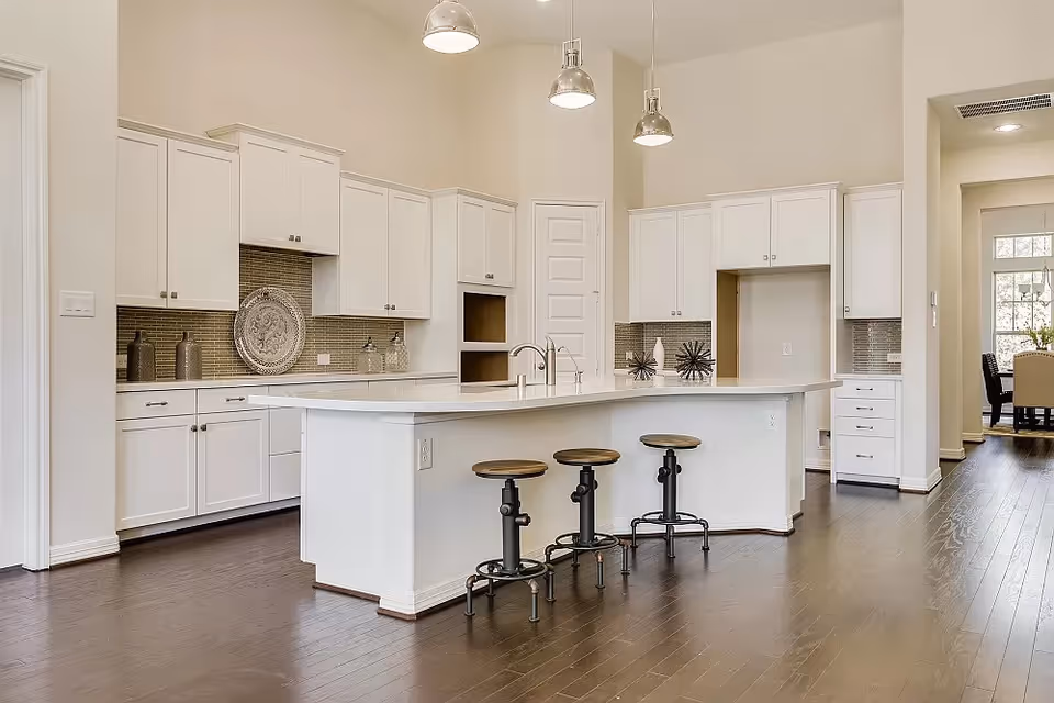 Bright modern kitchen with white cabinets, large island, pendant lights, and dark wood floors.