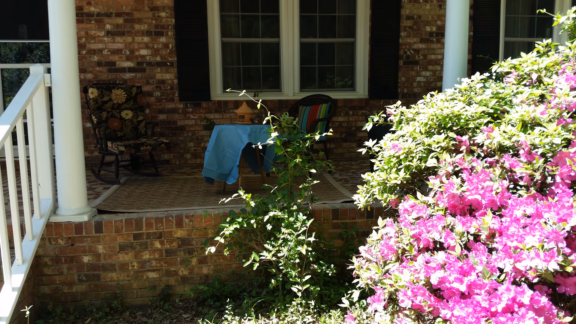 Brick porch with a small table, chairs, and vibrant pink flowering bushes.
