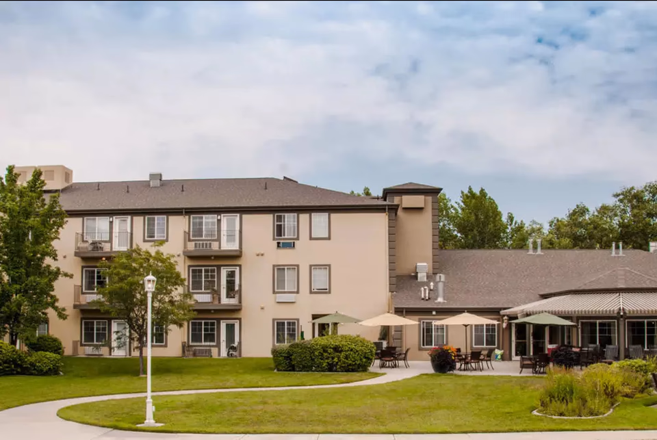 Exterior view of Cedarwood at Sandy, showing a three-story beige building with multiple windows and balconies, surrounded by green grass, trees, and outdoor seating areas with umbrellas under a partly cloudy sky.