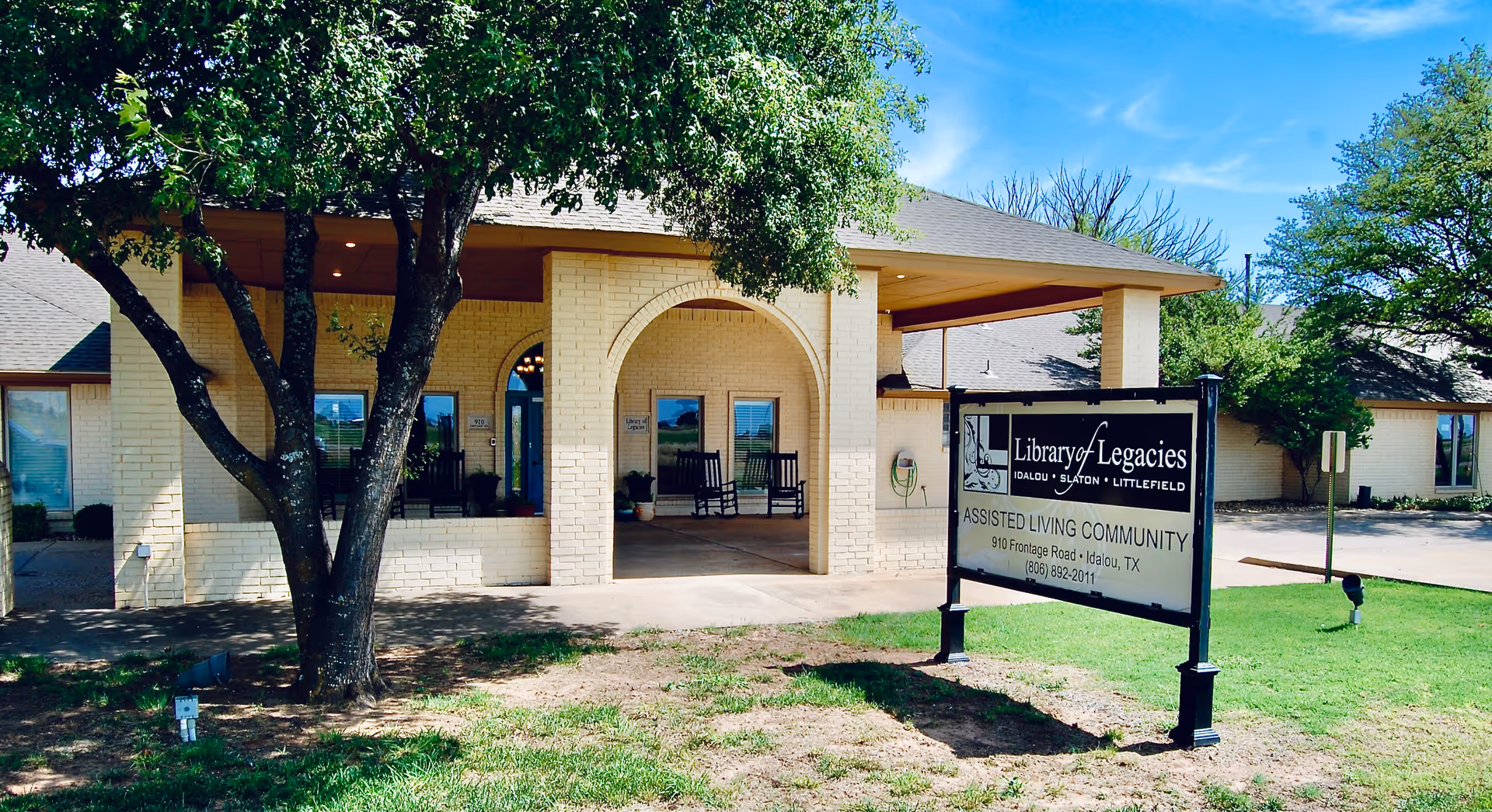 Exterior view of the Library of Legacies assisted living community building in Idalou, Texas, showing a light-colored brick facade with an arched entrance, rocking chairs on the porch, a tree in the foreground, and a sign with the facility name and contact information on the lawn.