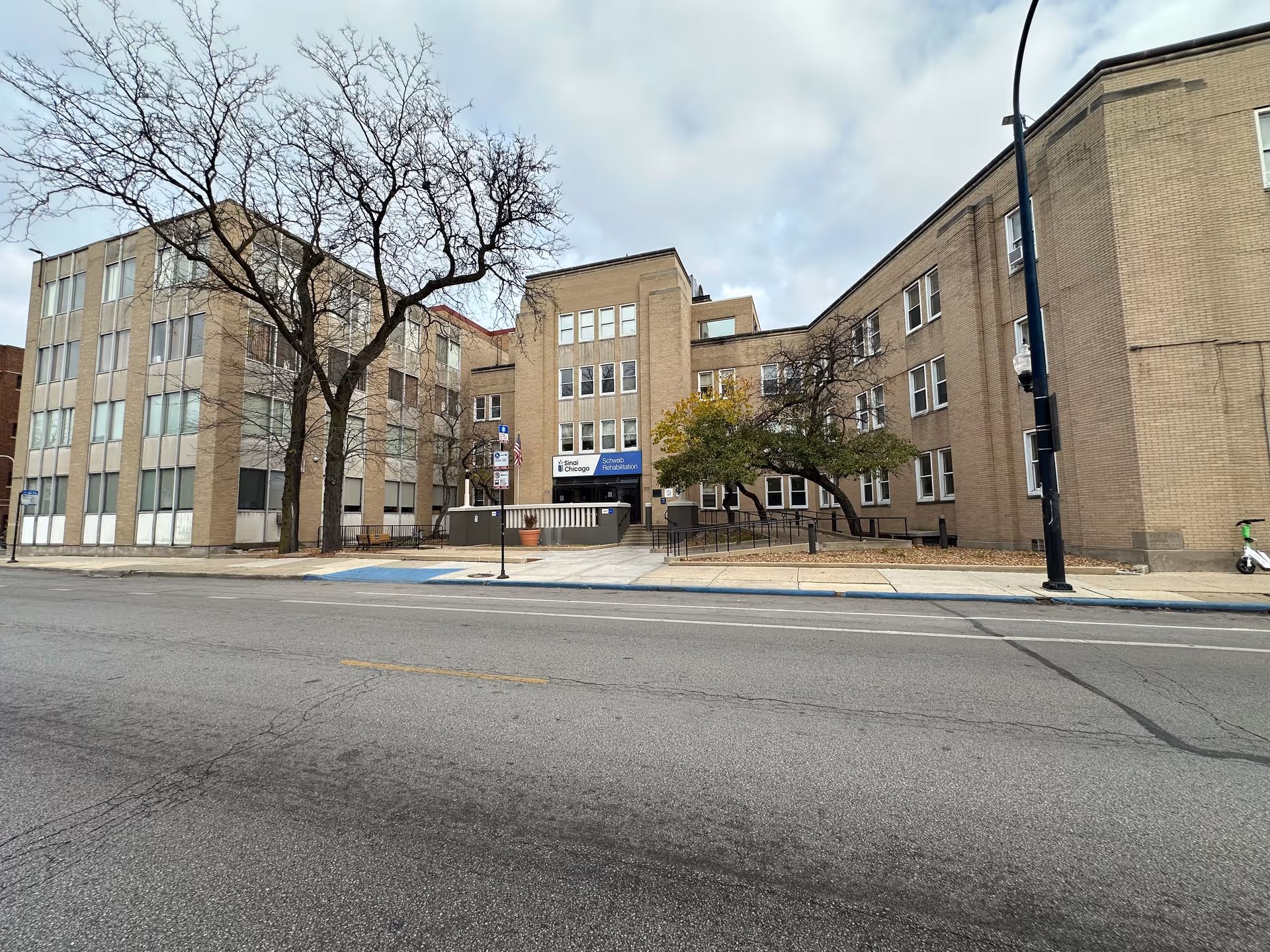 Street-level view of the front of a multi-story brick rehabilitation center building with bare trees and a sidewalk ramp.