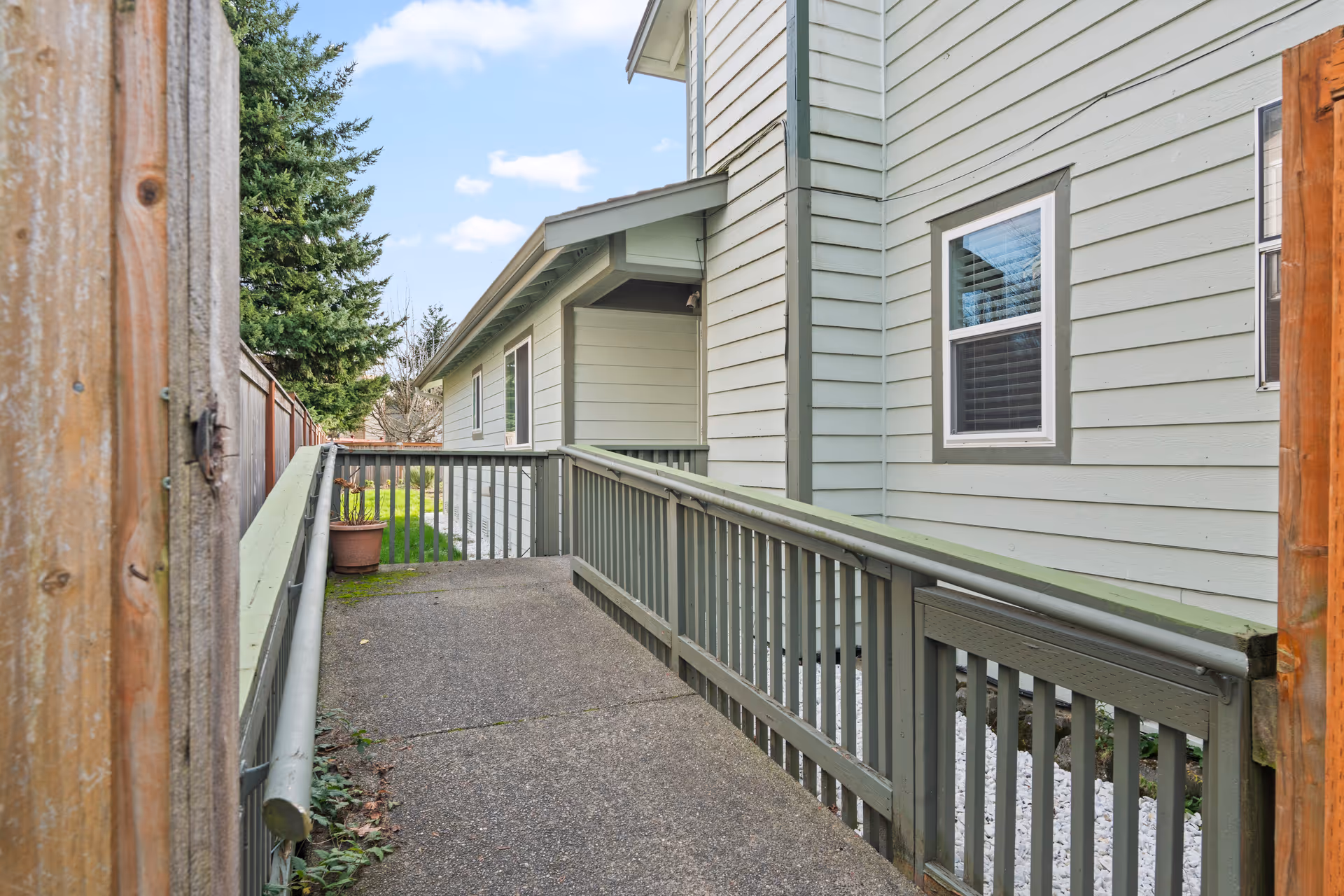 Exterior walkway beside a pale gray house with green railing and wooden fence.