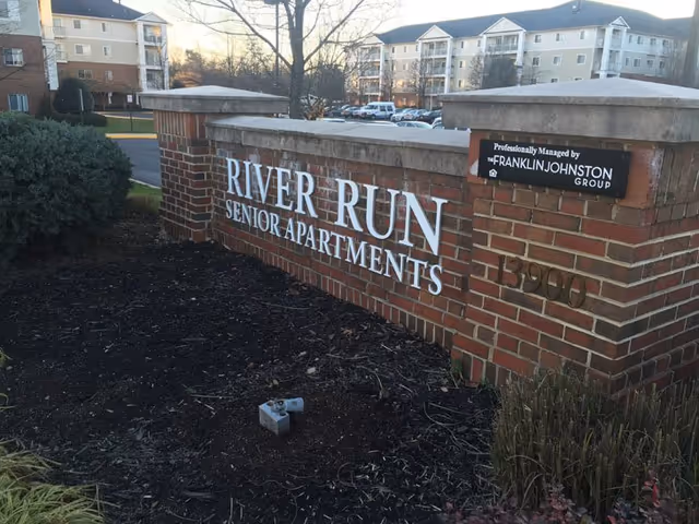 Brick entry sign reads River Run Senior Apartments with surrounding residential buildings in background.