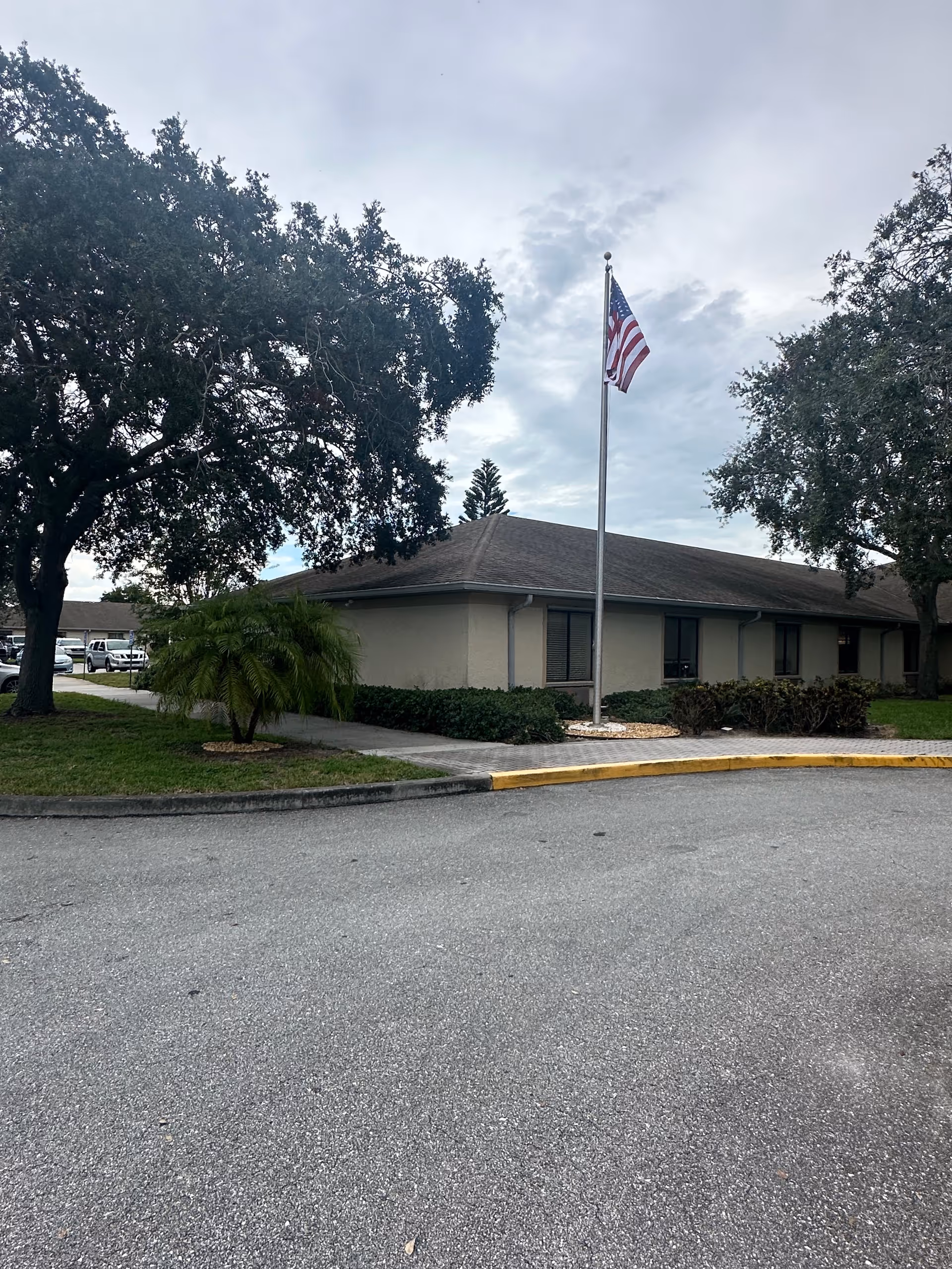 Single-story facility exterior with flagpole, American flag, large trees, and manicured landscaping.
