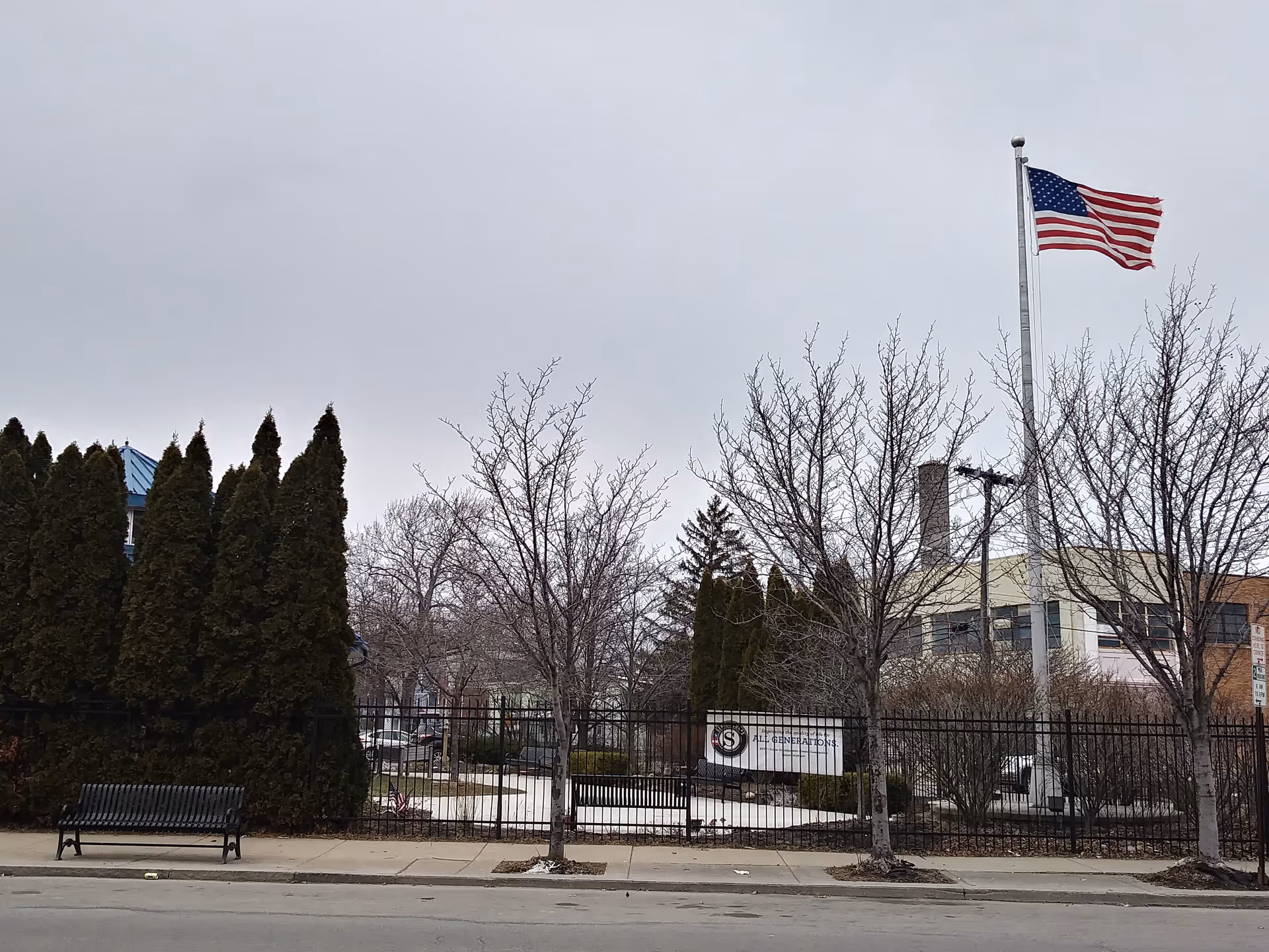 Exterior view of a senior care facility with American flag, bare trees, fence, and bench.