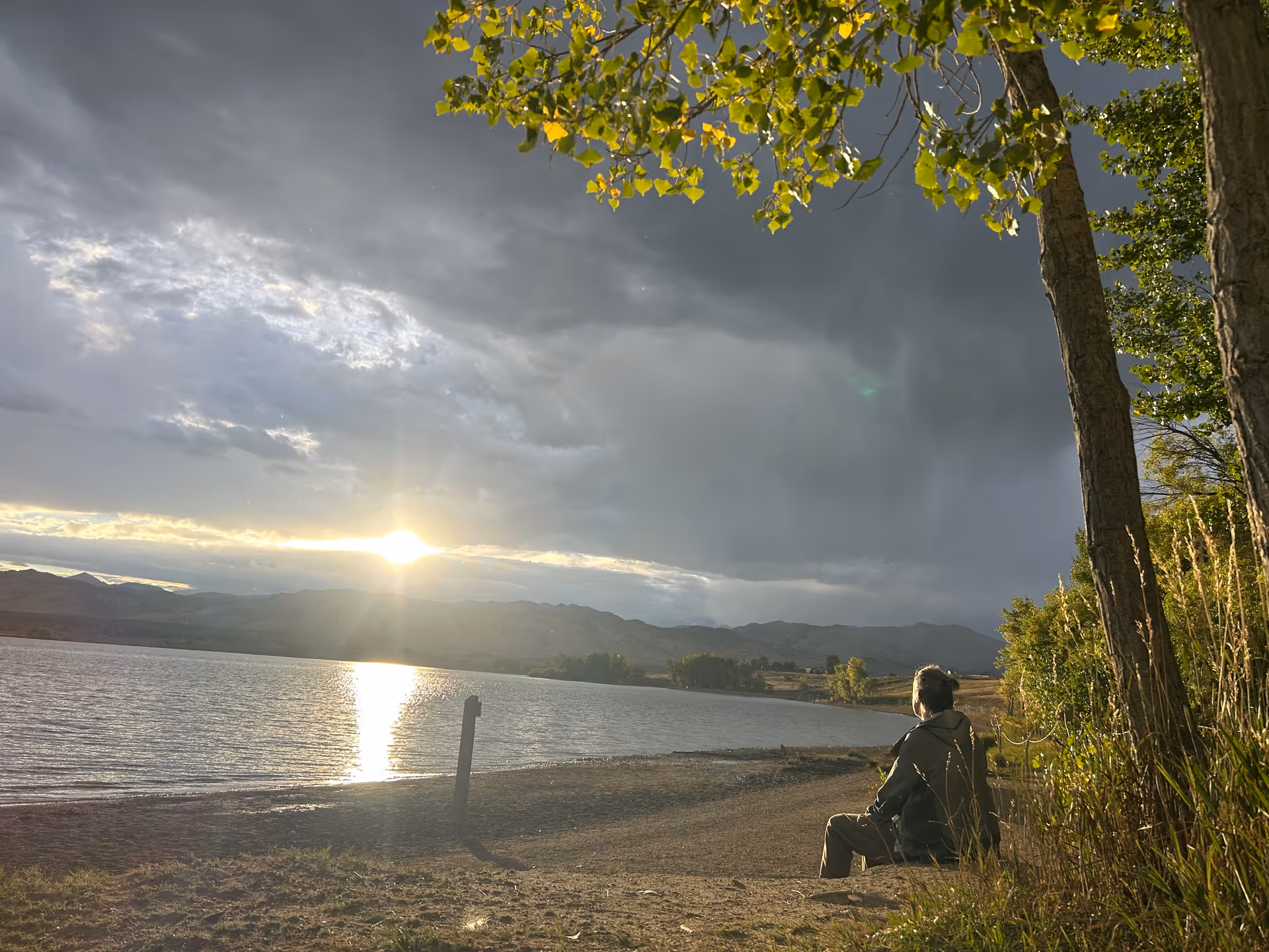 A person sits on a lakeshore at sunset, with trees and a dramatic cloudy sky.