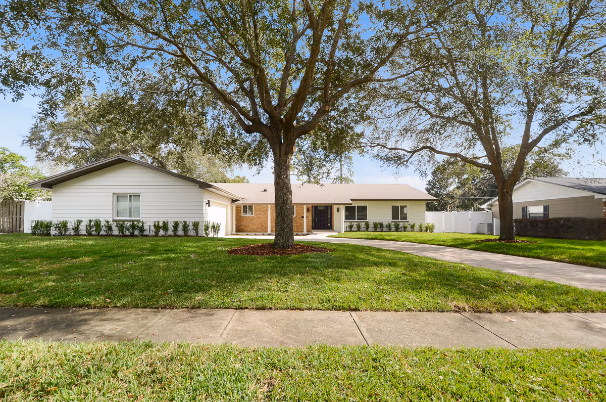 Single-story white ranch-style home with a large front lawn, two mature trees, and a curved driveway leading to the entrance.