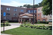 Brick-assisted living facility with a central covered entrance, manicured lawn, and shrubs.