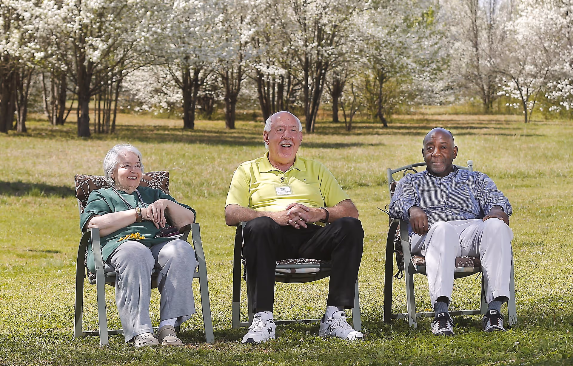 Three elderly individuals sitting on outdoor chairs on a grassy area with blooming trees in the background, enjoying a sunny day.