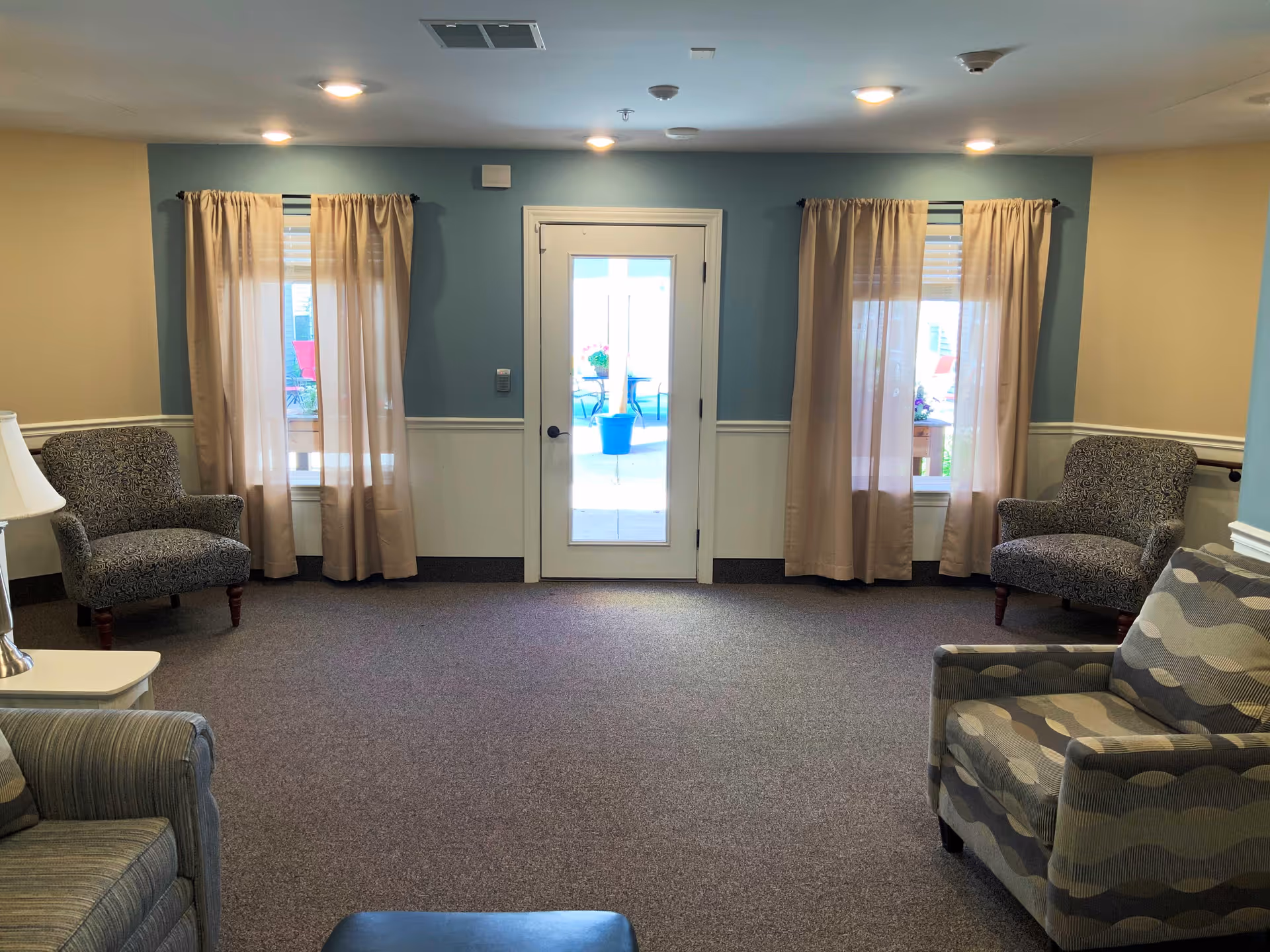 Carpeted common room with upholstered chairs arranged around a central space and a glass door flanked by curtained windows.