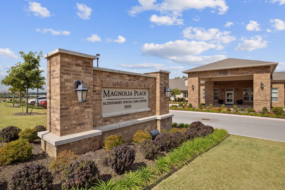 Exterior view of Magnolia Place Alzheimer's Special Care Center showing a brick sign with the facility name and address, surrounded by landscaped bushes and grass, with the building entrance visible in the background under a partly cloudy sky.