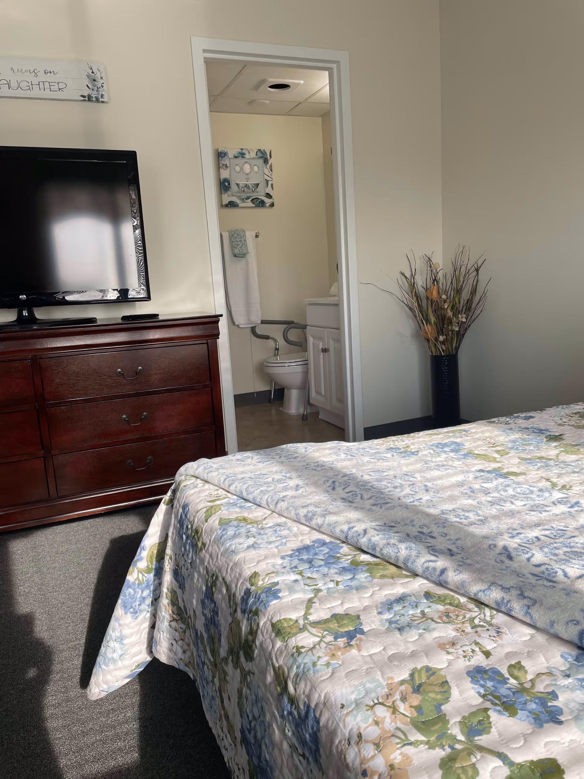 A bedroom with a floral bedspread in shades of blue and green. There is a dark wooden dresser with a flat-screen TV on top. An open door reveals a bathroom with a toilet, a white cabinet, a towel rack with a white towel, and a decorative wall hanging. A tall vase with dried branches is placed in the corner of the room.