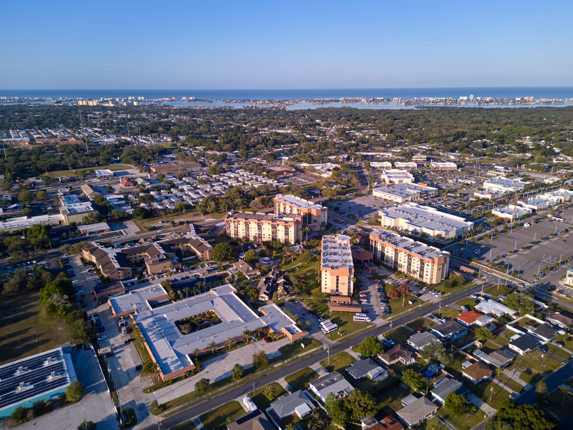 Aerial view of a senior living campus with multiple apartment buildings, surrounding neighborhood streets, and the coastline visible on the horizon.