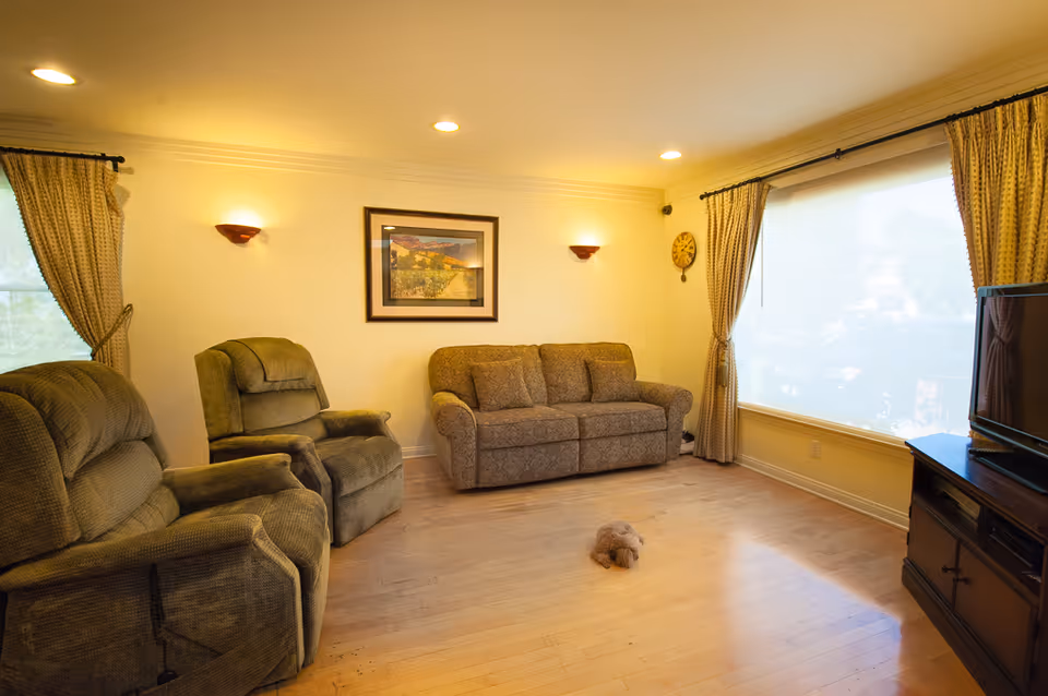 A cozy living room with two recliner chairs and a patterned sofa against a cream-colored wall. The room has wooden flooring, a large window with beige curtains, a wall clock, a framed landscape painting, and a TV on a wooden stand. A small stuffed animal is on the floor in the center of the room.