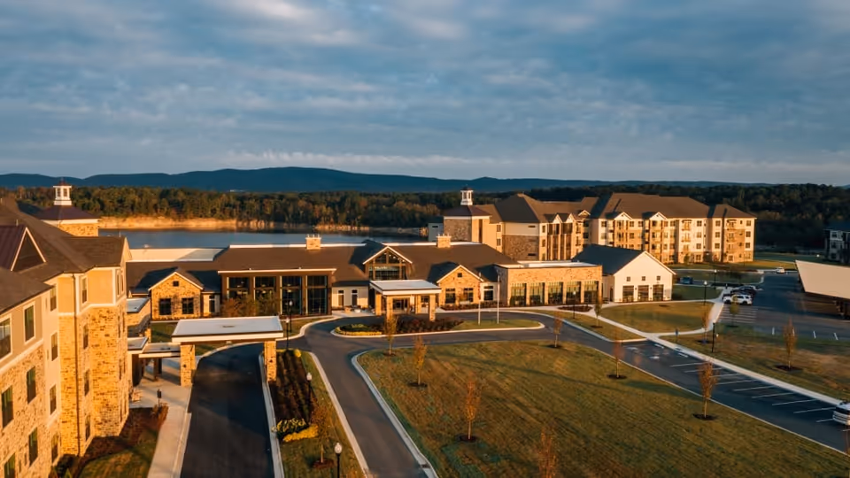 Aerial view of a senior living complex with its main entrance, driveways, parking lots, and a lake in the background.