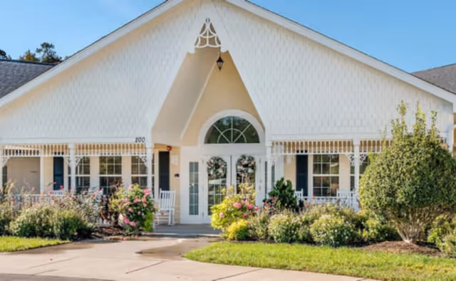 White gabled senior living facility entrance with shrubs and colorful flowers.