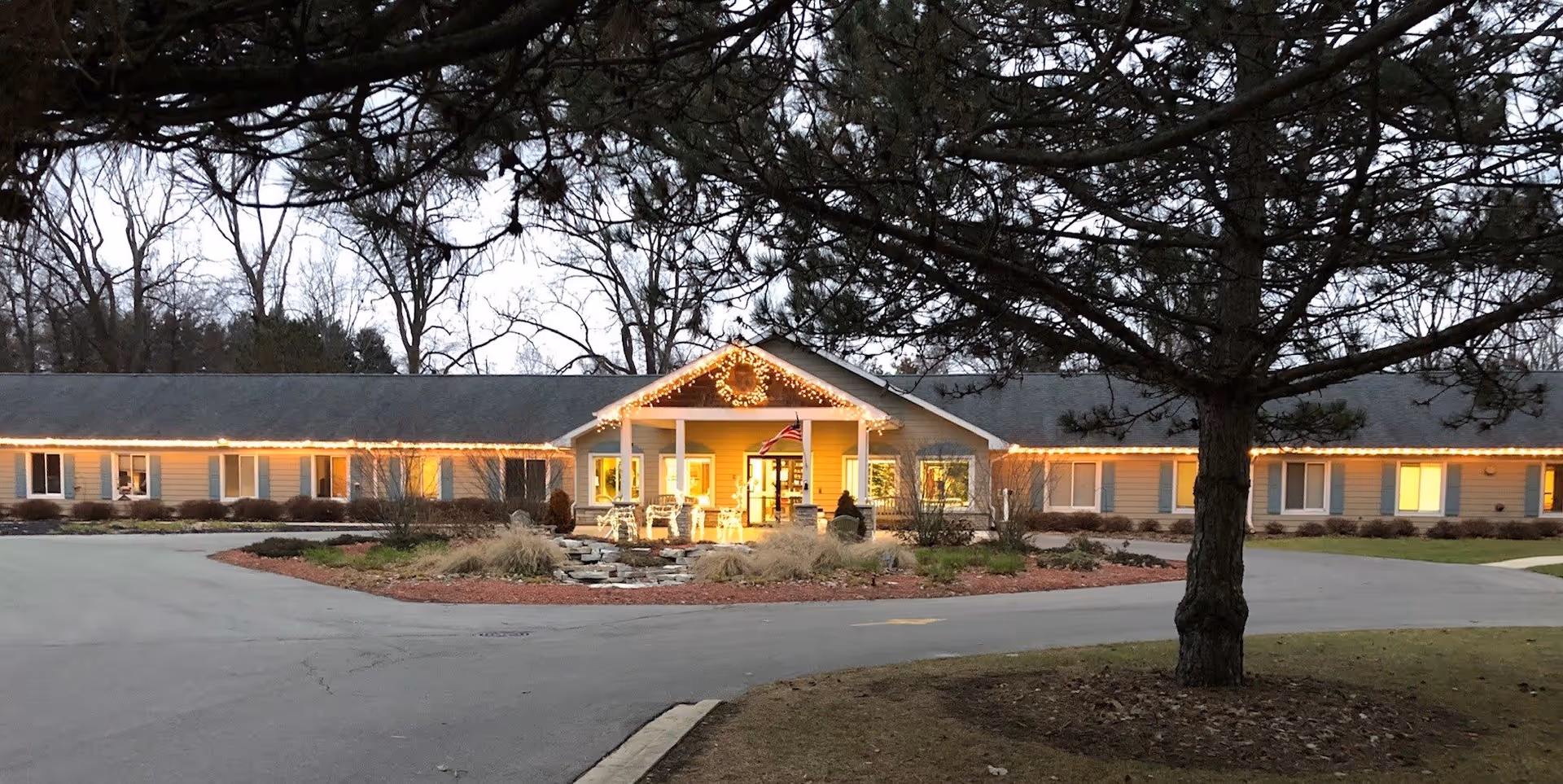 Front exterior of a single-story senior living facility lit with string lights at dusk, showing the entrance, circular driveway, landscaping, and a large tree in the foreground.