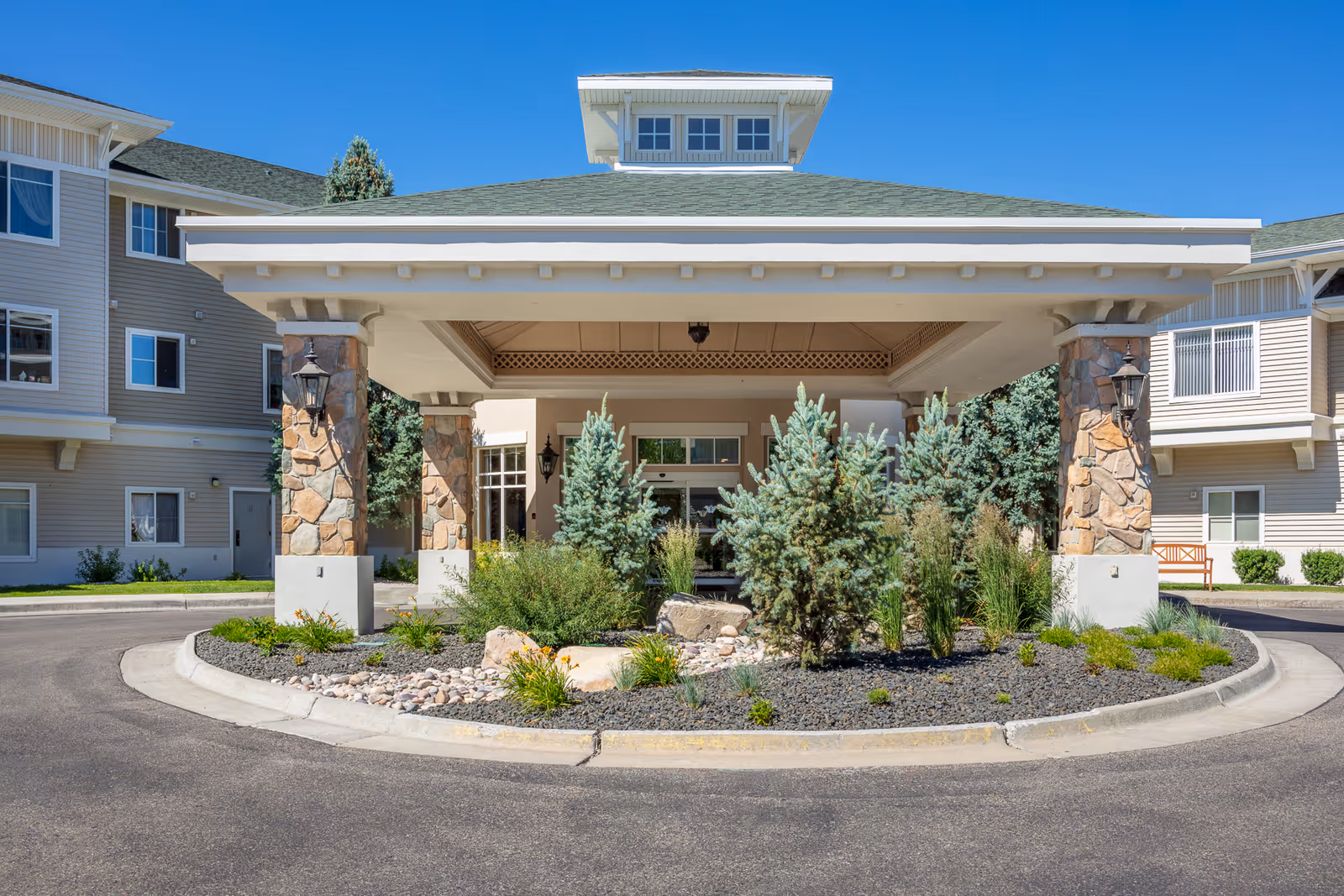 Entrance of a senior living facility with a covered drop-off area supported by stone pillars, surrounded by landscaped greenery and small trees, with the building visible in the background under a clear blue sky.