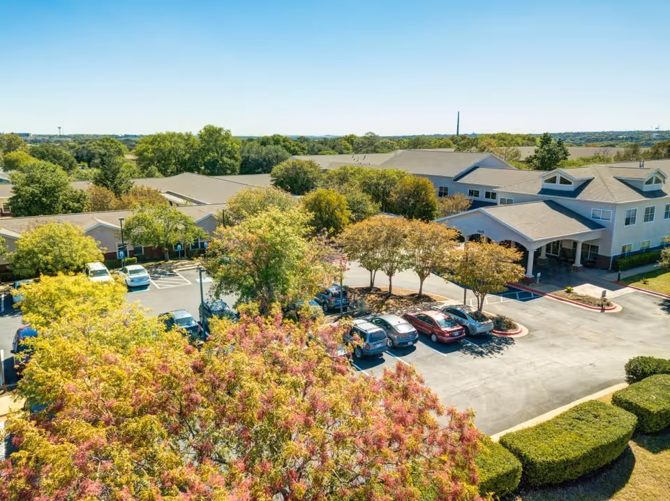 Aerial view of Parmer Woods at North Austin senior living facility showing the main building with a covered entrance, parking lot with several cars, and surrounding trees with green and reddish foliage under a clear blue sky.