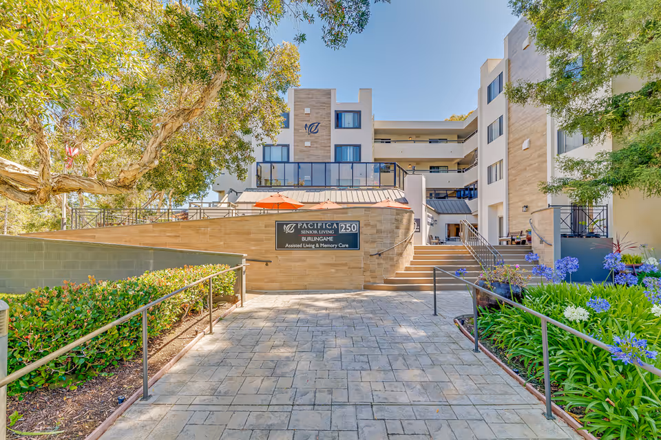 Front entrance of the Burlingame senior living building with a paved walkway, ramp and steps, landscaping, and a sign reading 'Pacifica Senior Living Burlingame'.