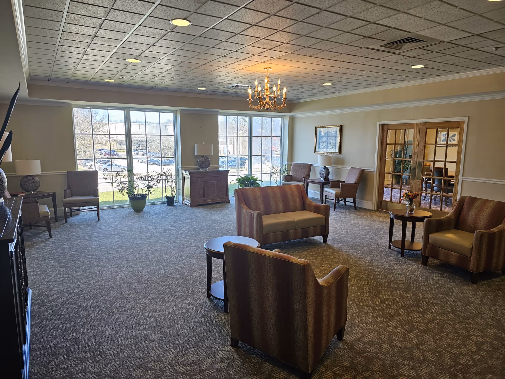 A spacious, well-lit living room area with large windows letting in natural light. The room features several upholstered chairs and loveseats arranged around small round tables. There are potted plants near the windows, table lamps on side tables, a chandelier hanging from the ceiling, and framed artwork on the walls. Through glass-paneled double doors, another room with additional seating is visible.