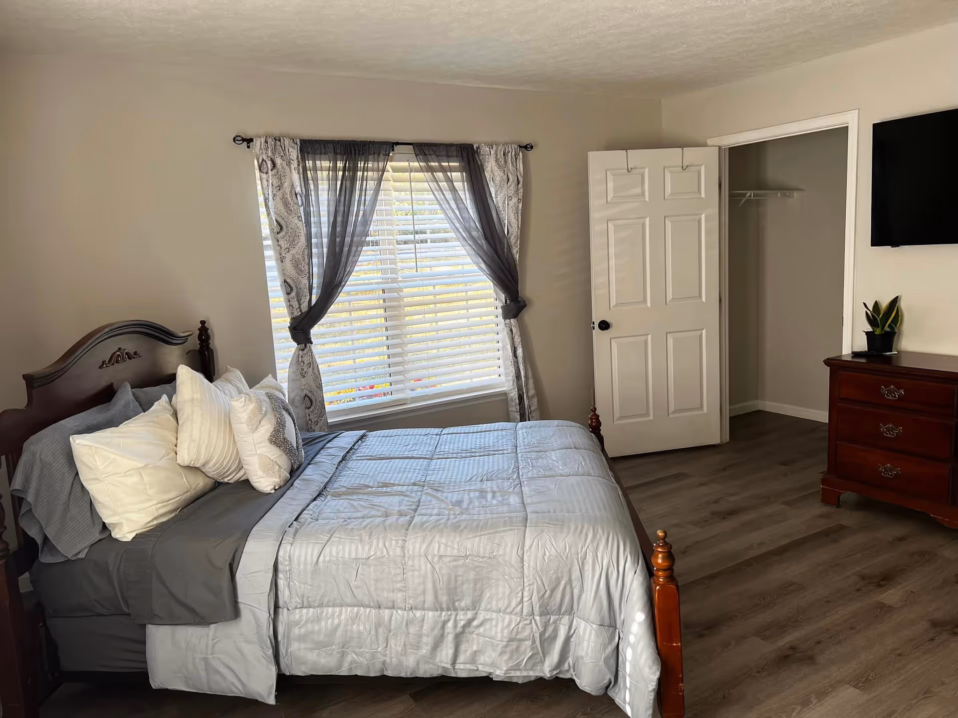 Bedroom with a wooden bed, gray bedding, window with sheer curtains, dresser and closet.
